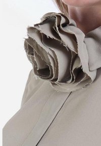 Blusa in tessuto beige con un grande fiore in tessuto texturizzato come accentuazione al collo. Il fiore presenta bordi stratificati e sfrangiati e curve morbide.