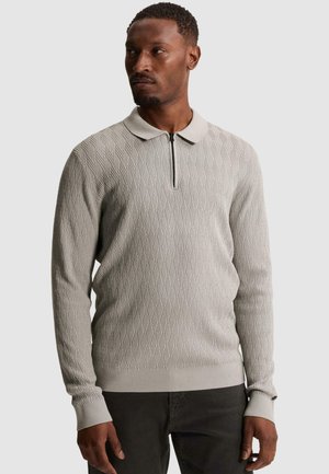 Άνδρας που φοράει ανοιχτό γκρι υφαντό πουλόβερ με μακριά μανίκια, κοντό φερμουάρ και γιακά, κοιτάζει ελαφρώς στο πλάι.