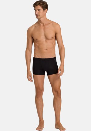 Man standing barefoot wearing black fitted boxer briefs, looking to his left against a plain light background.