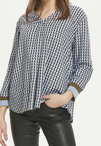 Kvinna som bär en långärmad mönstrad blus med blå, vita och bruna geometriska former, tillsammans med svarta läderbyxor.