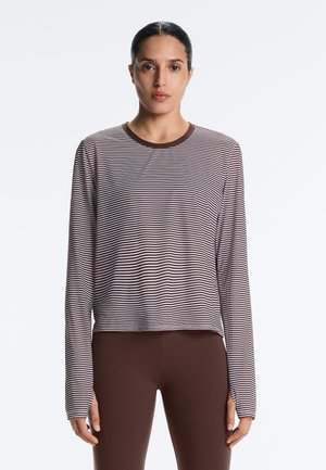 Žena odjevena u smeđu i bijelu prugastu majicu dugih rukava s otvorima za palce i jednobojne smeđe helanke, stoji nasuprot jednobojnoj pozadini.