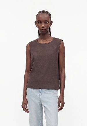 Žena s vrkočmi, oblečená v hnedý tielko a svetlomodré rifle, stojaca na jednoduchom bielom pozadí.