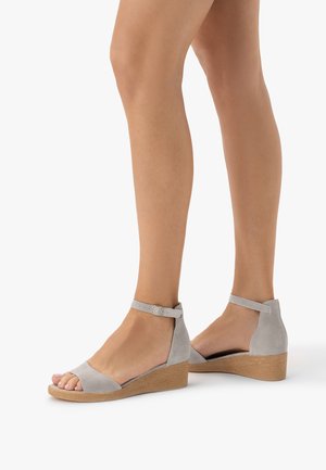 Nogi w jasno szarych zamszowych sandałach na koturnie z paskiem wokół kostki oraz beżowymi teksturowanymi podeszwami na białym tle.