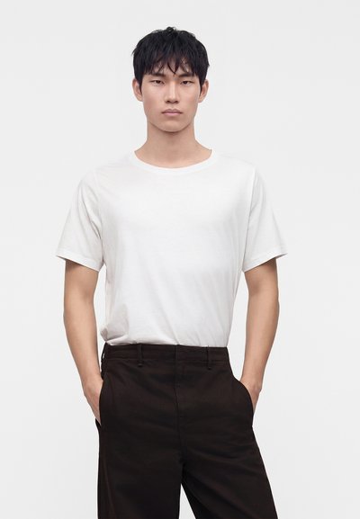 Jeune homme debout face à l'avant, portant un t-shirt blanc uni à manches courtes et un pantalon noir, les mains dans les poches, sur fond blanc.