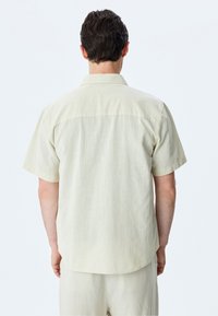 Camisa de manga corta beige con una tela texturizada, dobladillo recto y un cuello sencillo, que presenta un diseño limpio y minimalista.