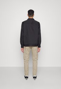 Man met zijn rug naar de camera, gekleed in een zwarte jas, beige cargobroek met zijzakken, witte sokken en zwarte lage sneakers.