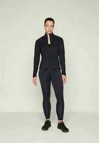 Joven mujer de pie, vistiendo una chaqueta negra con cremallera, leggings negros y zapatillas deportivas negras, contra un fondo claro y liso.