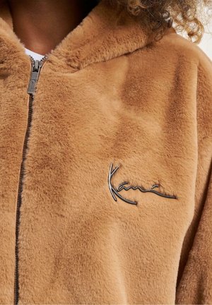 Beiger Kapuzenpullover aus Kunstpelz mit Reißverschluss, einer weichen Textur und einem metallischen Logoakzent auf der Vorderseite.