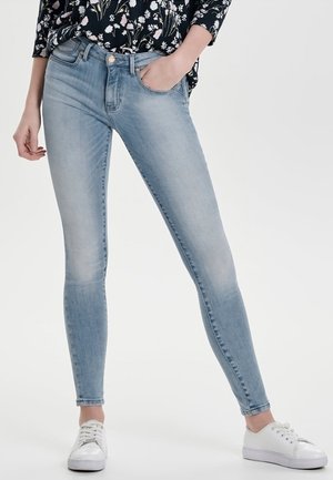 Woman wearing light blue skinny jeans, one hand in pocket, floral long-sleeve top, and white sneakers, standing against plain background.