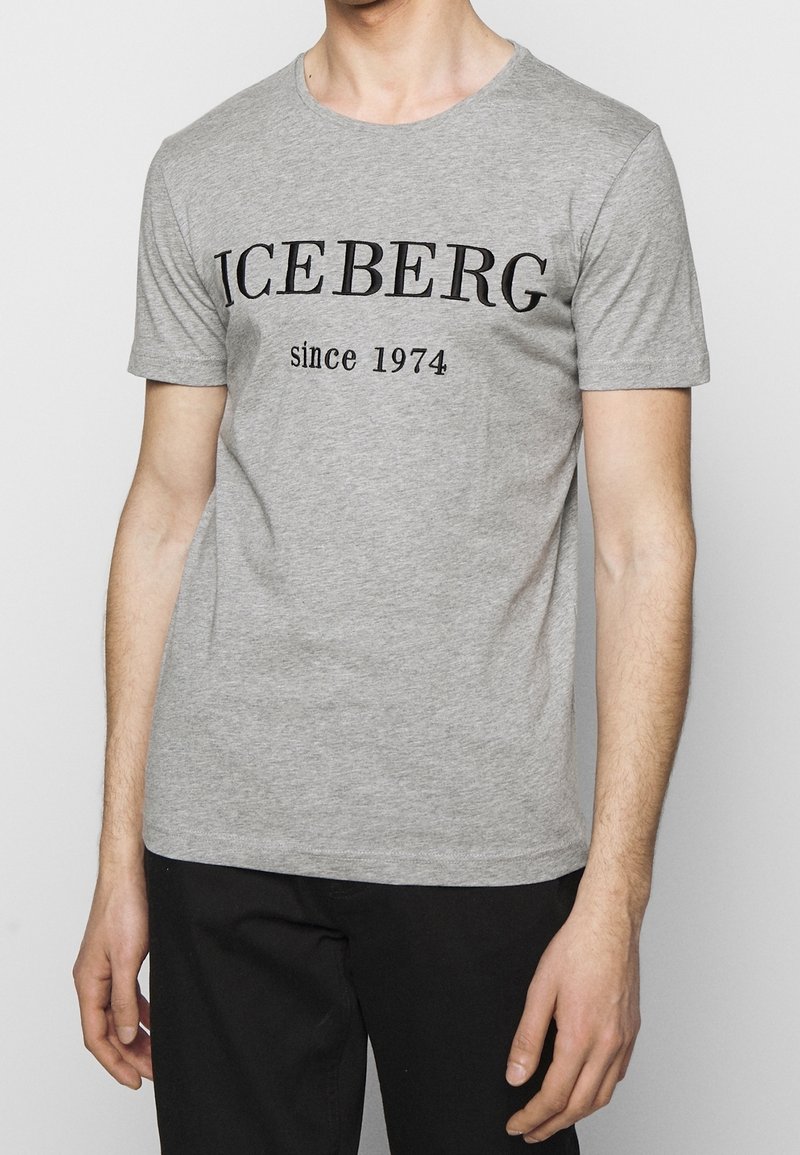 Homme portant un t-shirt gris à manches courtes avec l'inscription "ICEBERG depuis 1974" en noir sur le devant, associé à un pantalon noir.