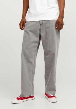 Mężczyzna ubrany w luźne szare jeansy z denimu, białą koszulkę i czerwone niskie trampki stoi na jednolitym tle.