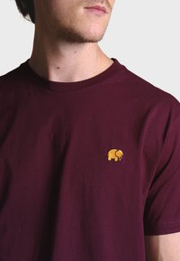 Burgundské bavlněné tričko s malým zlatě vyšívaným slonem na levé hrudi. Kulatý výstřih, krátké rukávy, hladká textura.