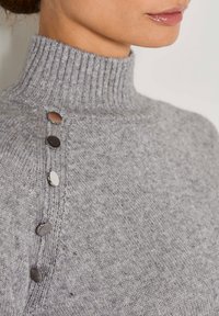 Frau, die einen grauen Strickrollkragenpullover mit fünf metallischen Knöpfen entlang der Schulternaht trägt.