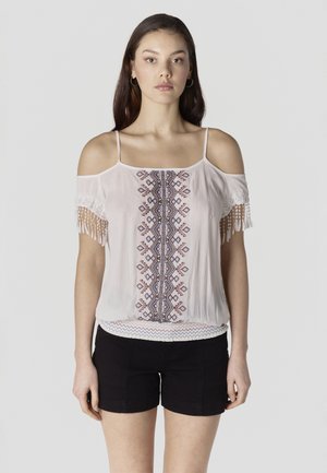 Joven mujer con una blusa blanca de hombros descubiertos con bordados geométricos y pantalones cortos negros, posando frente a un fondo liso.