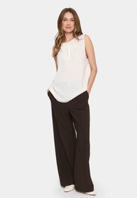 Blusa bianca senza maniche con scollatura arricciata e silhouette morbida, abbinata a pantaloni larghi di un marrone scuro, creando un contrasto di colori.