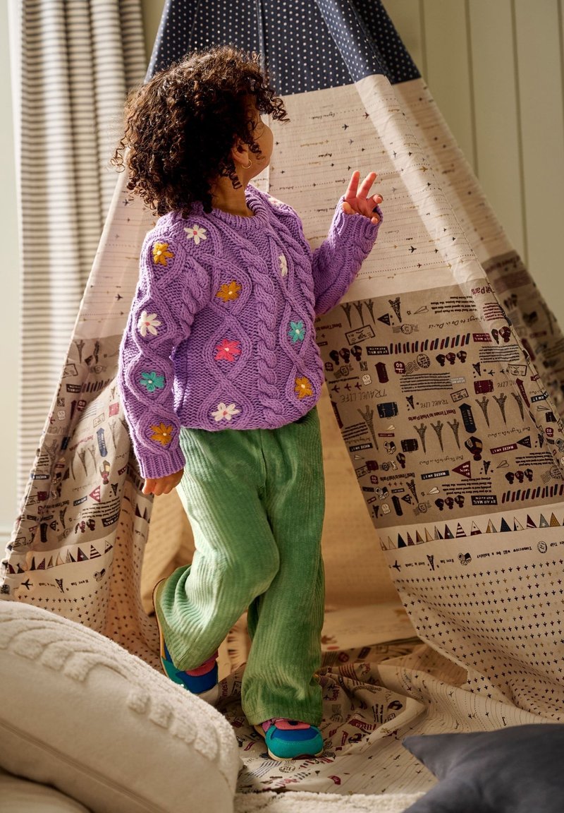 Enfant aux cheveux bouclés portant un pull violet à fleurs et un pantalon vert à l'intérieur d'une tente de jeu en tissu à motifs dans une pièce confortable.