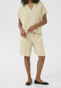 Set di abbigliamento da casa beige chiaro composto da un top loose-fitting con scollatura a V e maniche corte, abbinato a pantaloni corti fino a metà coscia. Scarpe nere piatte.