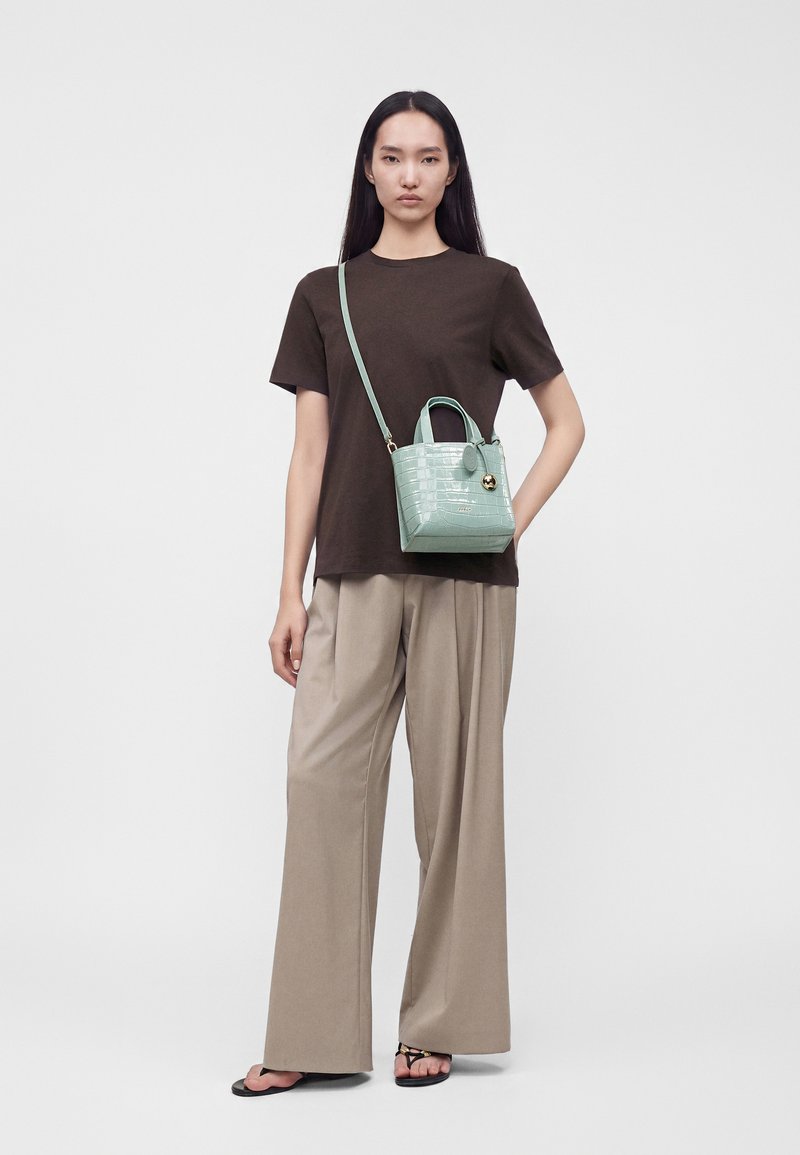 Femme portant un t-shirt marron et un pantalon large beige, tenant un petit sac bandoulière vert menthe avec un motif texturé et un accent doré.