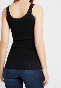 Frau in einem ärmellosen Top mit schwarzem Spitzenrand und blauen Jeans, von hinten vor einem schlichten weißen Hintergrund betrachtet.