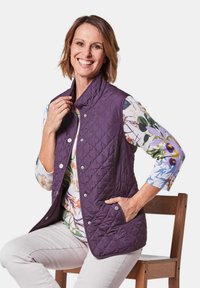 Gestepptes lila Weste mit hohem Kragen, Druckknöpfen und seitlichen Taschen, getragen über einem floral gemusterten Langarmoberteil.