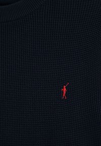 Dunkelblauer strukturierter Stoff mit einer kleinen roten, gestickten Figur, die auf der rechten mittleren Seite einen Polo-Schläger hält.