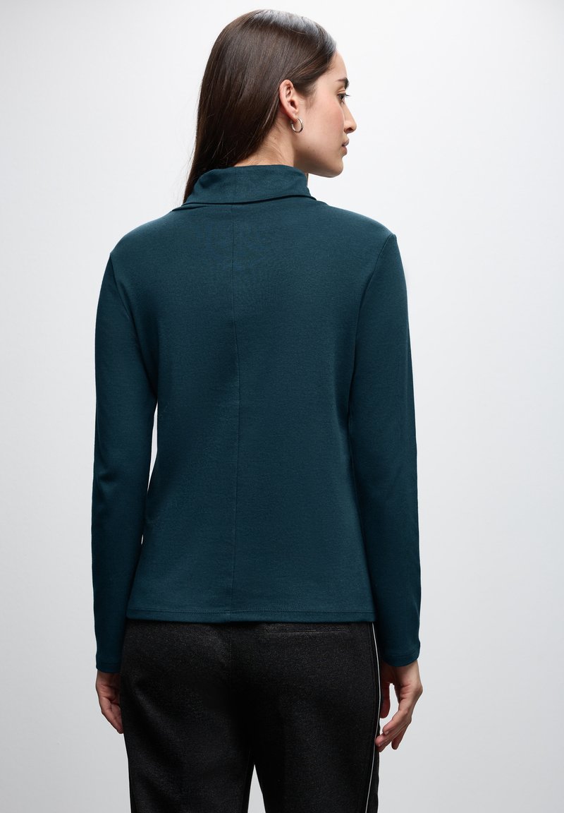 Longsleeve-Oberteil in dunkelblaugrün mit weicher Textur, hohem Kragen und subtilen Nahtdetails hinten, kombiniert mit dunklen Hosen.