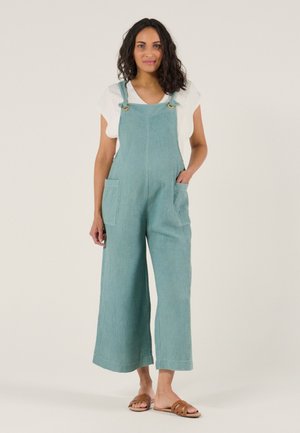 Frau trägt hellblaue, strukturierte, weit geschnittene Latzhosen und ein weißes T-Shirt, steht barfuß in braunen Pantoletten vor einfachem Hintergrund.