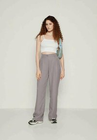 Young woman with curly hair wearing white crop top, gray high-waist pants, sneakers, and a light blue shoulder bag, standing against plain background.