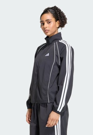 Jonge vrouw met krullend haar in een hoge paardenstaart, draagt een zwart sportjack en broek met witte strepen, kijkt naar de camera.