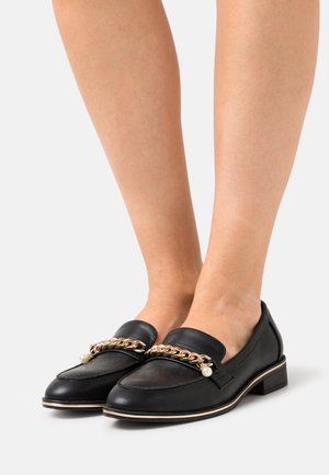Mocassins en cuir noir à petit talon carré, avec un accent en chaîne dorée et un petit détail en perle à l'avant. Texture lisse, bout rond.