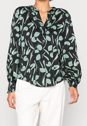 Blouse noire avec un motif abstrait de feuilles vertes et blanches, manches longues bouffantes avec poignets froncés, portée avec un pantalon blanc.