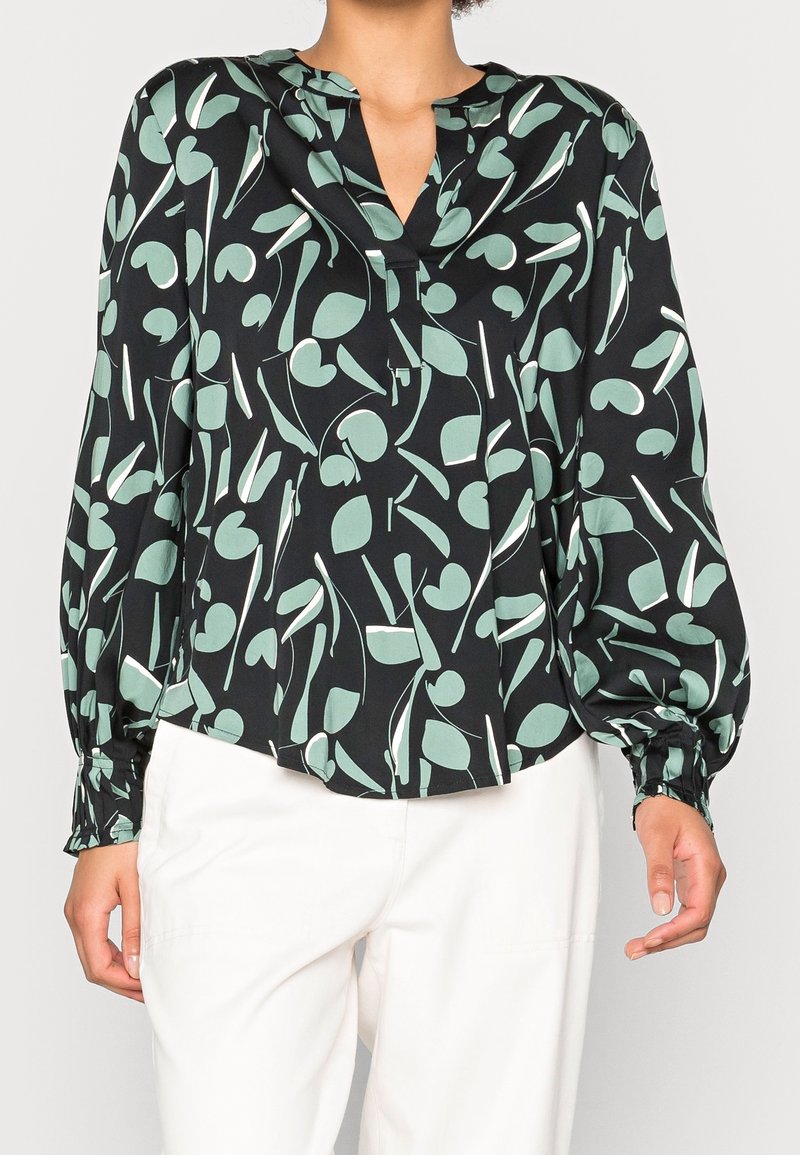 Blouse noire avec un motif abstrait de feuilles vertes et blanches, manches longues bouffantes avec poignets froncés, portée avec un pantalon blanc.