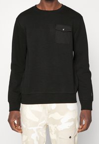 Svart crewneck sweatshirt i mjukt tyg, med en enda bröstficka med tryckknapp och ribbade ärmslut. Bärs med kamouflagebyxor.