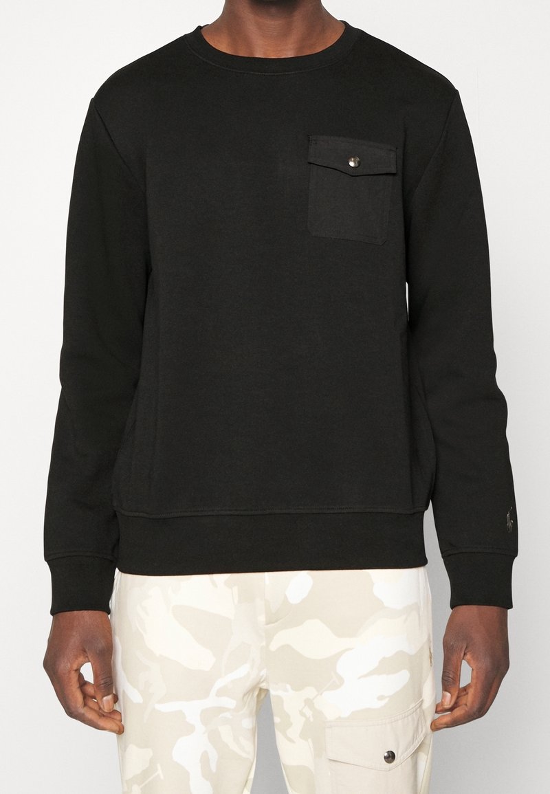 Svart crewneck sweatshirt i mjukt tyg, med en enda bröstficka med tryckknapp och ribbade ärmslut. Bärs med kamouflagebyxor.