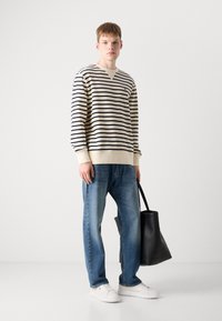 Stripet genser i krem og marineblå, løs passform; kombinert med lyseblå jeans og hvite sneakers. Modellen holder en svart totebag.
