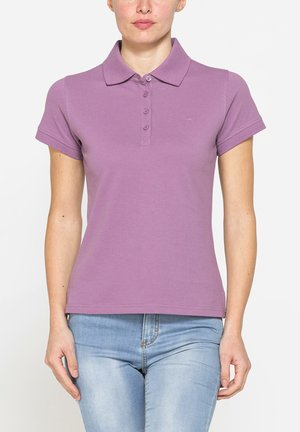 Donna che indossa una polo lavanda a maniche corte con bottoni abbinata a jeans azzurri, in piedi davanti a uno sfondo semplice.