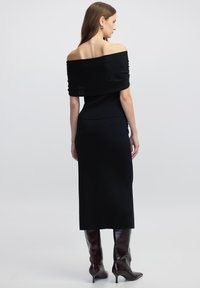 Zwarte off-the-shoulder gebreide jurk met een nauwsluitend lijfje en een midi-lengtede rok. Glad textuur. Gecombineerd met hoge, glanzende bruine laarzen.