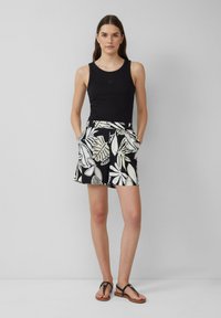 Top noir sans manches associé à un short à motifs de feuilles noir et blanc avec des poches latérales. Le mannequin porte des sandales plates, se tenant devant un fond clair.