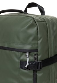 Mochila de tela texturizada verde que cuenta con un bolsillo con cremallera, correas negras y cierre de hebilla. Forma rectangular con bordes reforzados.