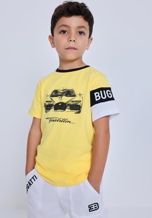 Ragazzo giovane che indossa una maglietta gialla Bugatti con uno schizzo di un'auto e pantaloncini bianchi con loghi Bugatti, in piedi davanti a uno sfondo semplice.