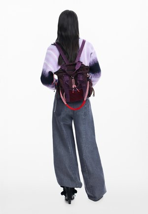 Persoană cu păr lung și închis la culoare, purtând un pulover cu gradient mov, blugi gri cu pantalonii largi, cizme negre și un rucsac mov cu bretele roșii și roz.