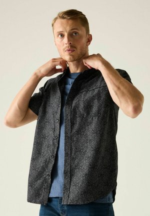 Mężczyzna poprawia kołnierz ciemnej, wzorzystej koszuli z krótkim rękawem, noszonej na niebieskim t-shircie i dżinsach, na jednolitym tle.