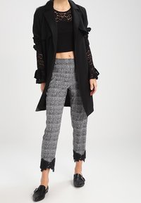 Top corto in pizzo nero sotto un trench coat nero, abbinato a pantaloni a quadri grigi con rifiniture in pizzo nero sull'orlo e mocassini neri.
