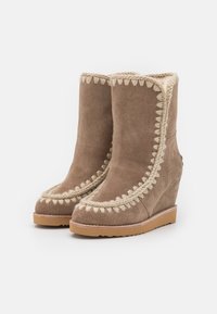 Botas de tacón cuña de ante marrón con un intrincado bordado en crema en la parte superior y los lados, y suela de goma texturizada.