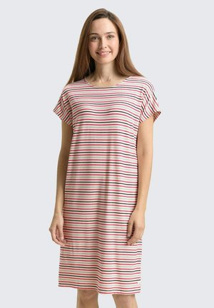 Femme debout et souriante, portant une robe mi-longue à manches courtes avec des rayures horizontales rouges, noires et blanches sur un fond clair.