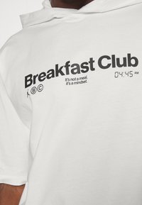 Witte hoodie met zwarte tekst die "Breakfast Club" luidt, met een digitale klok die "04:45 AM" weergeeft. Slogan inbegrepen.