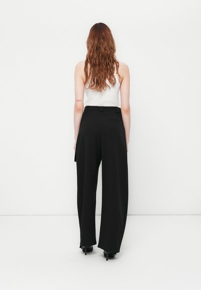 Femme aux longs cheveux roux portant un haut sans manches blanc et un pantalon large taille haute noir, se tenant devant un fond blanc.