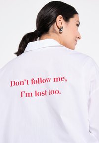 Camicia bianca con colletto classico e maniche corte. Sul retro è stampato un messaggio in rosso: "Non seguirmi, sono perso anch'io."