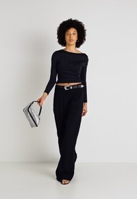 Melnas garas piedurknes topiņš ar pieguļošu dizainu, augstu apkakli un apgrieztiem malu, apvienots ar platām melnām biksēm un sudraba metālisku somu.