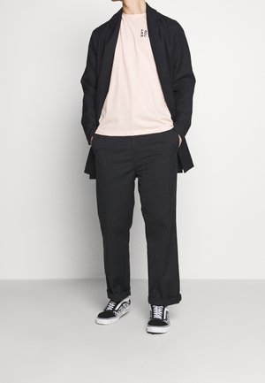 Abrigo largo negro, camiseta rosa con texto, pantalones negros y zapatos en blanco y negro. Diseño simple, ajuste relajado y aspecto casual.